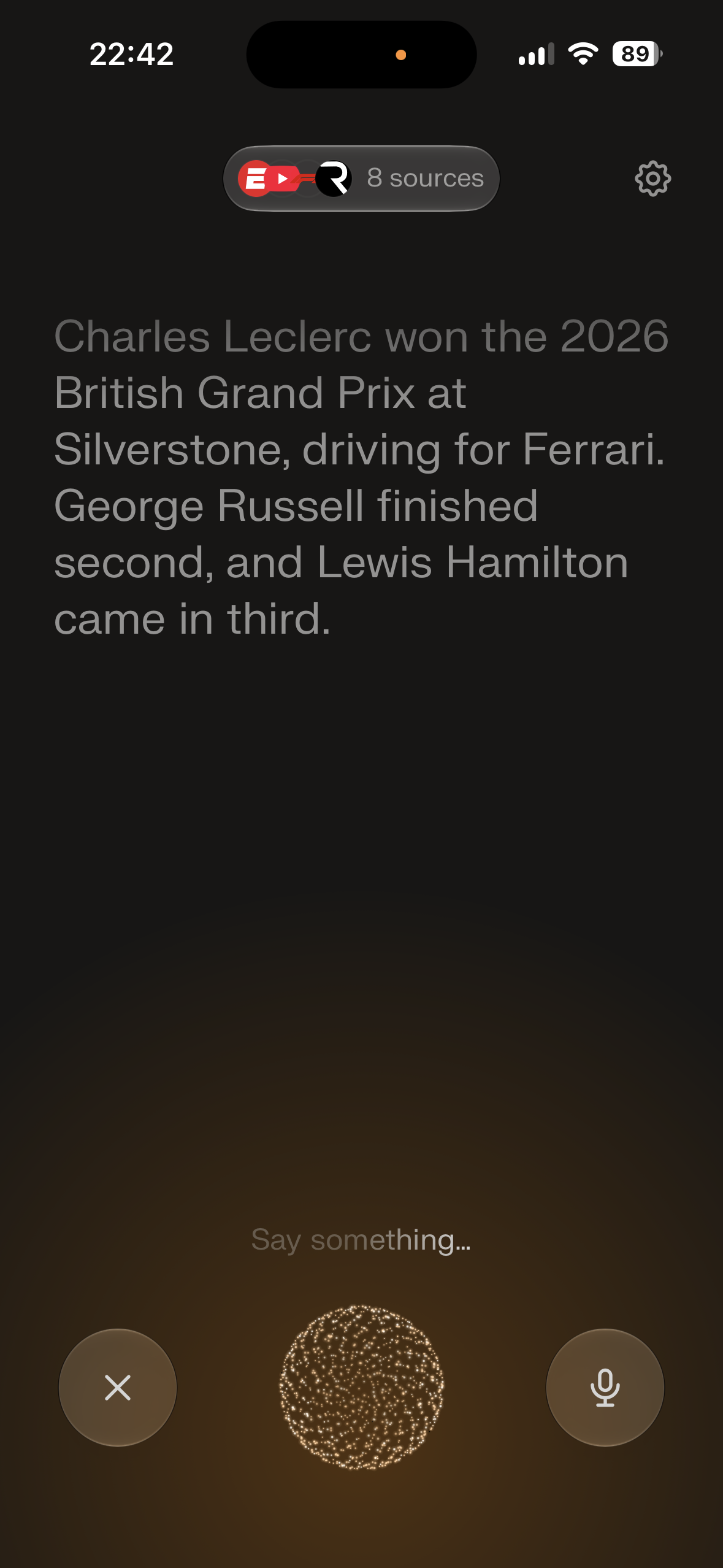 Dark mobile screen showing a spoken-answer interface with a large text response about the 2026 British Grand Prix, a sources pill near the top, a settings icon, and bottom controls for cancel and microphone with a pulsing orb in the center.